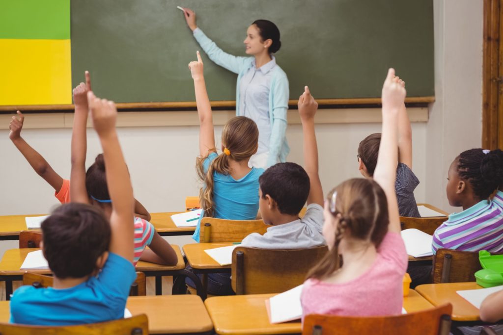 A teacher asks her elementary class a question and they all raise their hands.