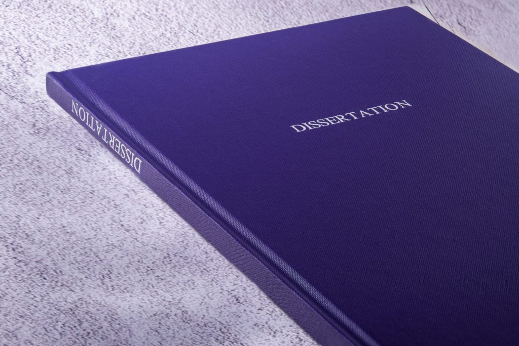 A closed blue book sits on a table with “dissertation” printed on the front cover.