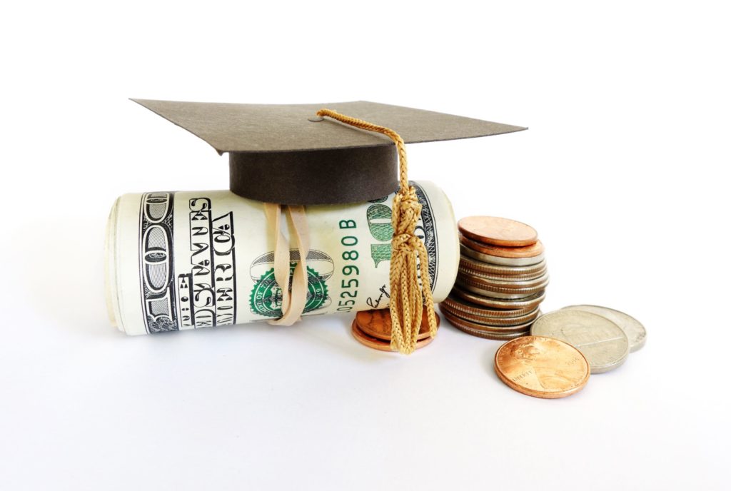 Graduation cap on top of wad of hundred dollar bills and stack of coins.