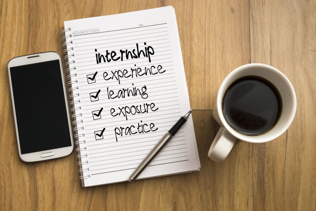 Benefits of an internship (experience, learning, exposure, practice) are written down on a notepad next to a cup of coffee.