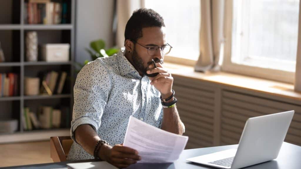 A man sits at his laptop with a paper in his hand, thinking.