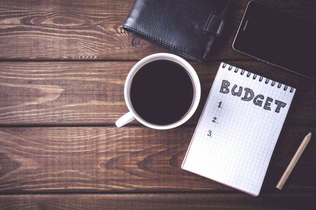 An overview of a notepad with a budget list, a wallet, phone, and cup of coffee.