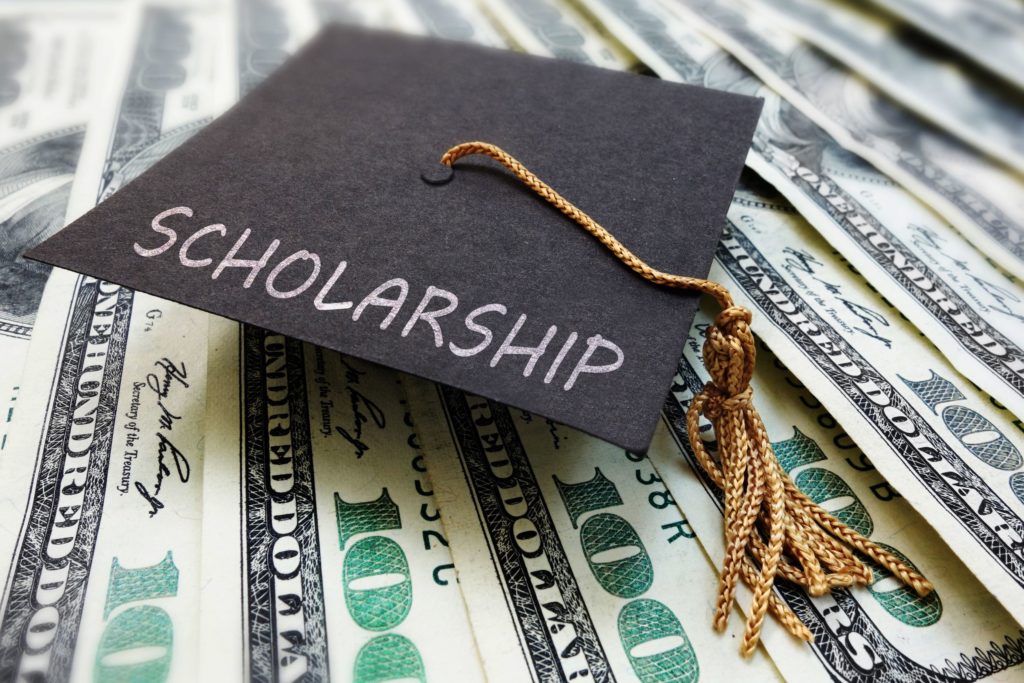 A graduation cap with the words “scholarship” sits on a pile of money.