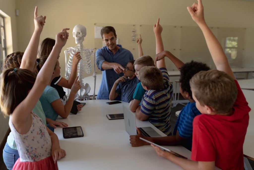 Teacher pointing to one student with his hand up surrounded by other classmates.