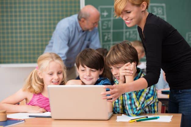 A smiling younger teacher helps a student while an older teacher helps students in the back of the classroom.